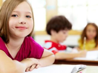 Une élève souriante en premier plan, vêtue d'un haut rose, assise à son bureau dans une salle de classe, tandis que d'autres enfants sont concentrés sur leur travail en arrière-plan, illustrant l'environnement d'apprentissage serein et engageant pour les enfants.